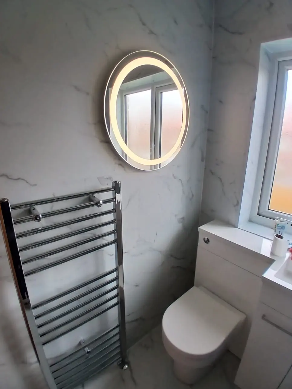 White vanity, back-to-wall toilet and chrome heated towel rail against white marble cladding in Jarrow